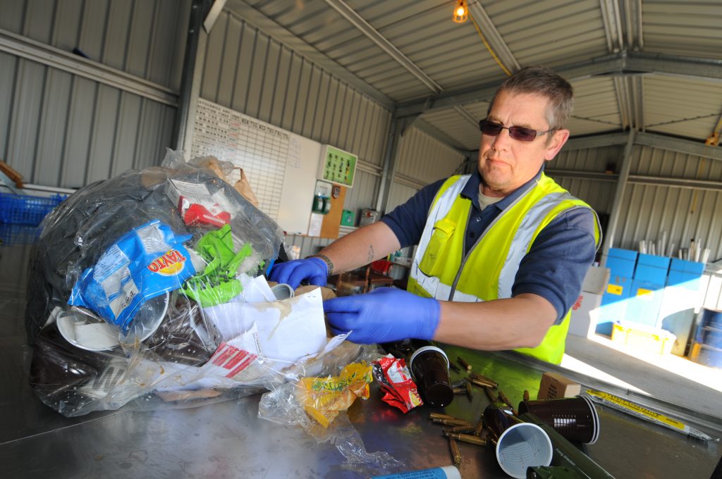 An employee from Landmarc wearing a hi-vis sleeveless jacket with blue gloves and brown glasses is putting away a clear recycling bag . They are some empty coffee cups beside the plastic bag and he is in a grey building with a whiteboard on the left-hand side and blue cabinets behind him.