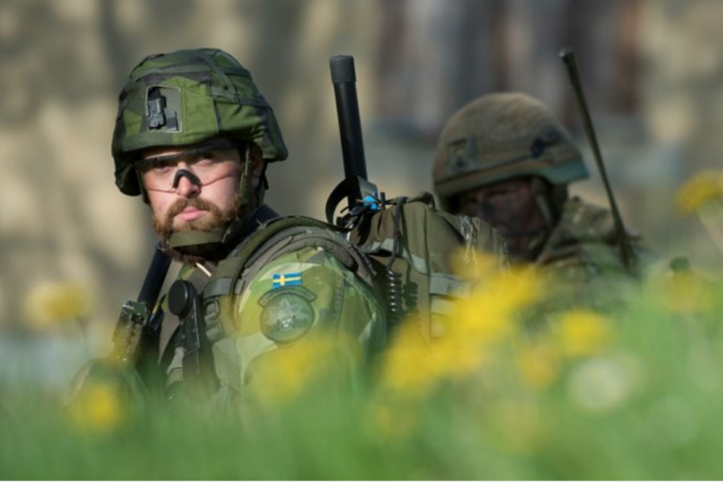 A Swedish soldier, in uniform and standing in profile, is looking towards the camera, with a British soldier behind him. 