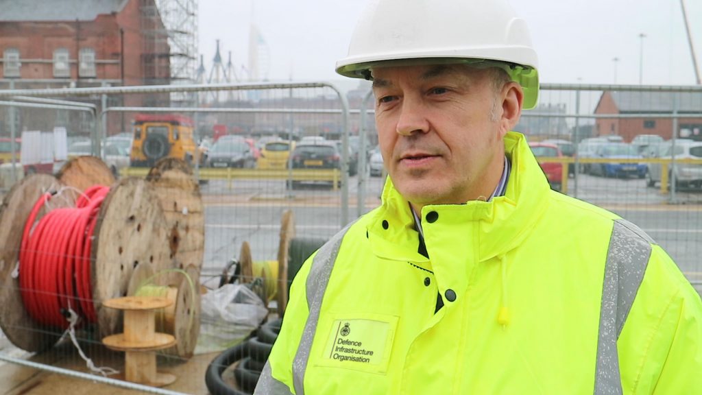 Project Manager, Colin Efford is pictured in a hi-vis jacket and helmet. Behind him is construction work taking place at HMNB Portsmouth