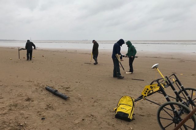 Four people on a beach, using equipment to find buried ordnance. In the foreground is a piece of scanning equipment on a wheel. 
