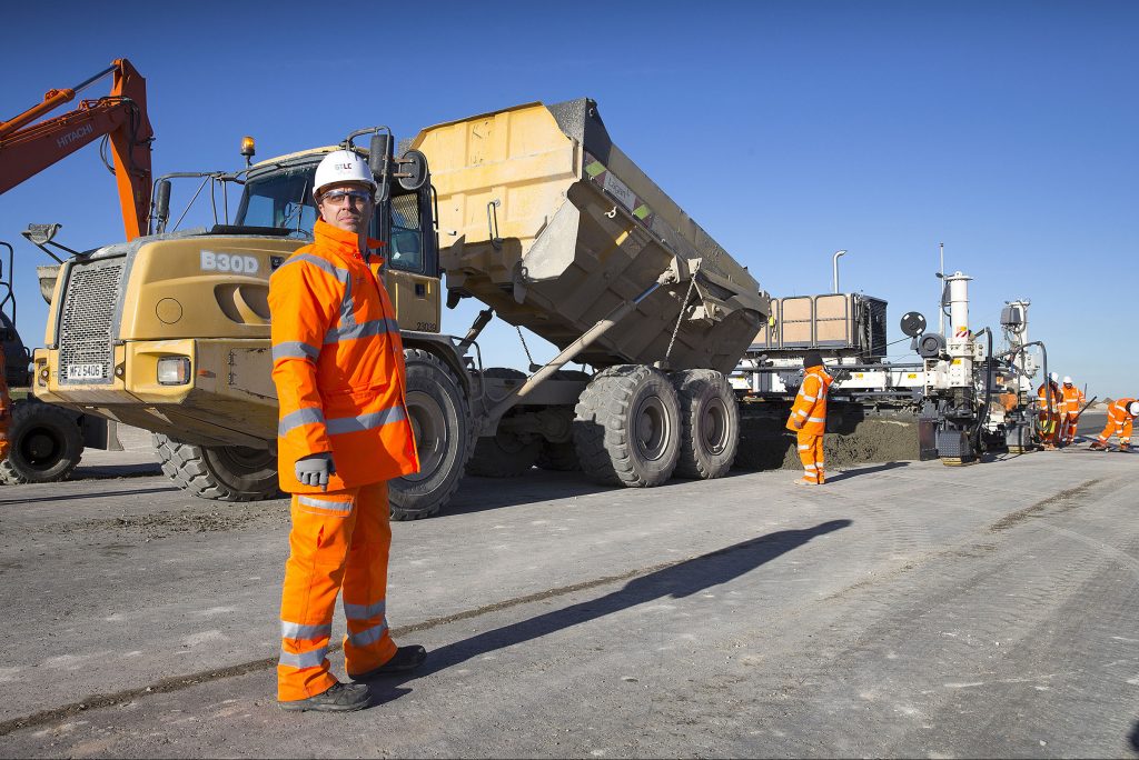 A construction worker dressed in orange high-vis and a protective helmet stands on the runway at RAF Marham, with a large vehicle behind. 