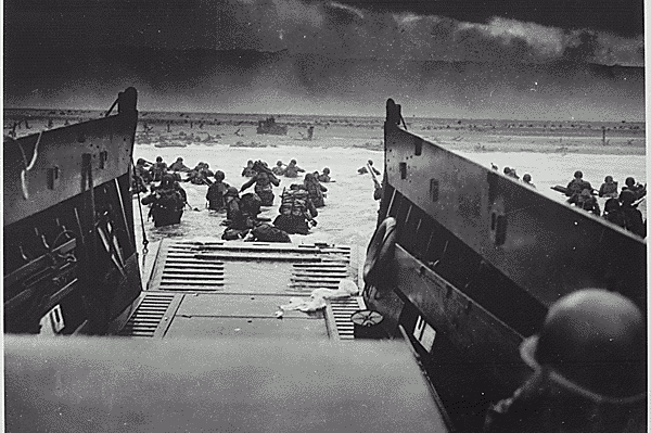 An original D-Day photo, taken from the inside of a landing craft. The black and white image shows a landing craft with the ramp down and soldiers wading through the sea towards the beach. 