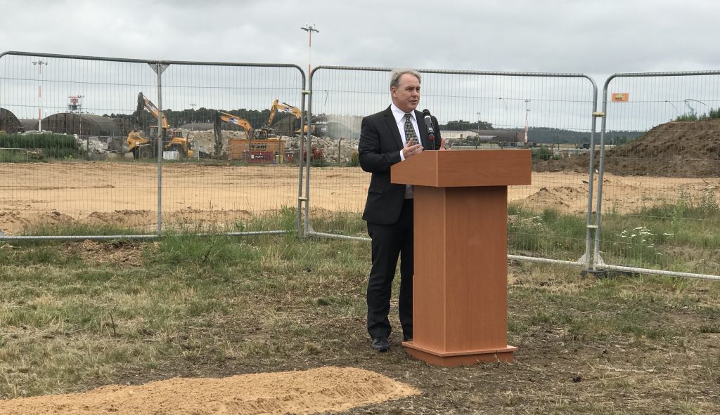 Keith Maddison, USVF Programme Director standing behind a wooden stand giving a talk about the importance of the facilities being delivered by DIO and its partners at RAF Lakenheath. Pictured are construction vehicles and a gate behind him.