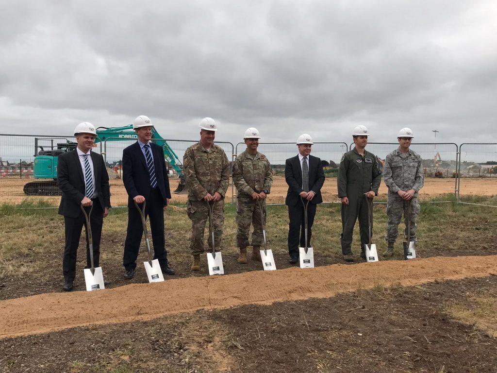 There are 7 people standing infront of a line of soil with a silver digger each and in hard hats to mark the start of construction at RAF Lakenheath. 
