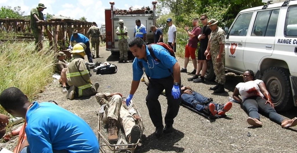 Pictured are the Belize Emergency Response Ambulance Service team and fire service responding to casulties in the exercise. A military vehicle is parked on the right-hand side of the road with a casulty next to it. A casulty is in the middle and a member of the emergency response team is responding to him. 
