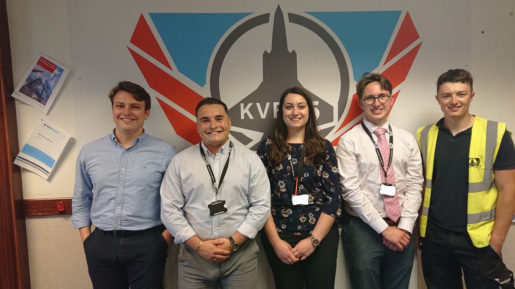 Pictured are 5 apprentices standing infront of a KVF35 logo that has a grey F35 with a circle around it and red wings. There are two men on the ledt in blue shirts, a girl in the middle with a black flowery top and two men on the right, one with a yellow hi-vis jacket.