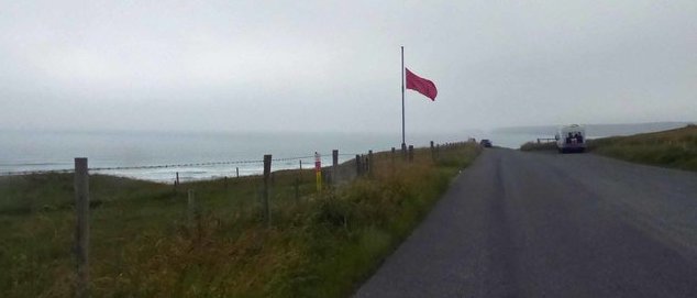 Pictured is Castlemartin Ranges with a path, greenery on the left-hand side, the sea and a red flag that is raised at the end.