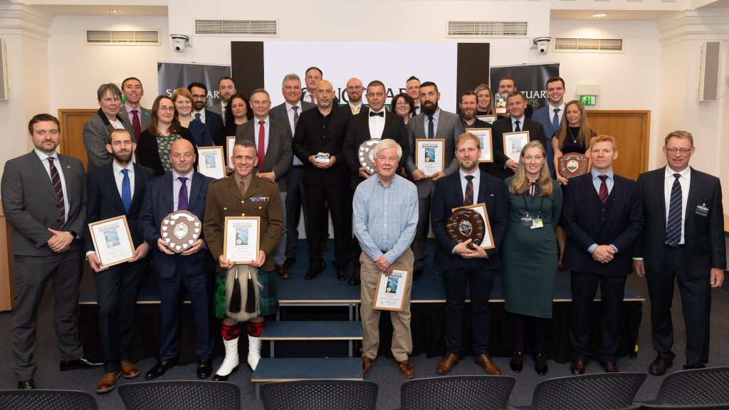 Pictured are a group of individuals from across the MOD in smartwear that have won the 5 categories of the Sanctuary Awards. They are standing on a blue stage with banners that say Sanctuary Awards behind them.