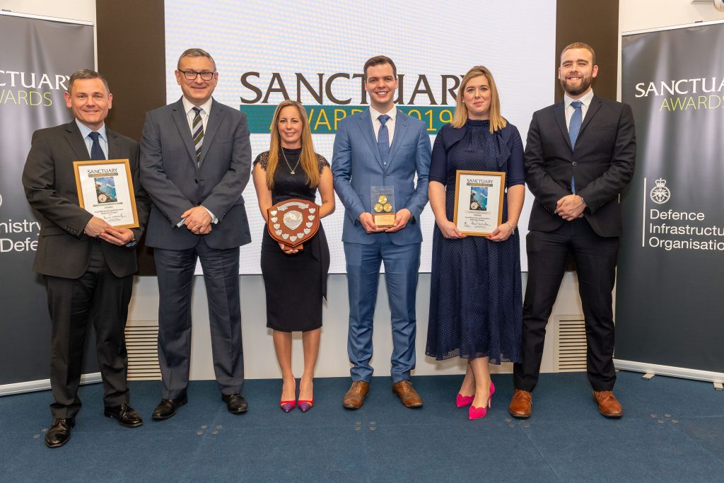 Pictured are 6 people standing on a blue stage with a screen behind them that says Sanctuary Awards. There is a man on the left in a suit and tie holding a framed award. In the middle is a women in a dress holding an award shaped like a shield. Next to her on the right is a man in a greay suit holding a clear award with three gold circles in it.