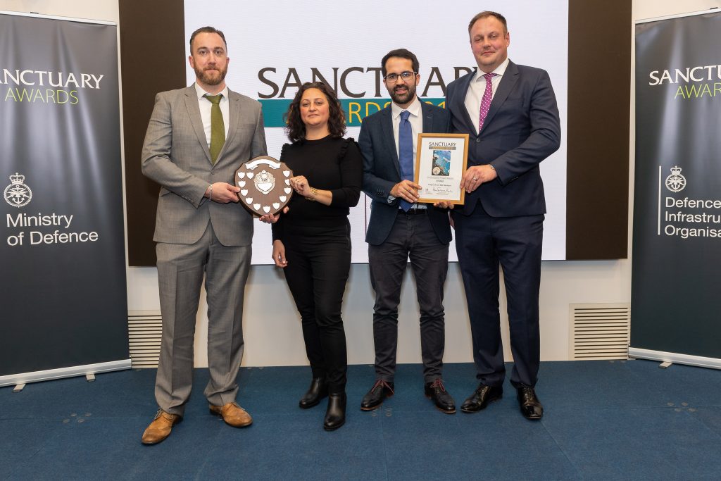 Pictured are 4 people standing on a blue stage with a screen in white and black and green writing that says Sanctuary Awards. The man on the left is wearing a greay suit and a lady next to him in black is holding an award shaped like a wooden shield with little silver shields around it. Next to the women on the right are to men in suits holding a framed wooden award.
