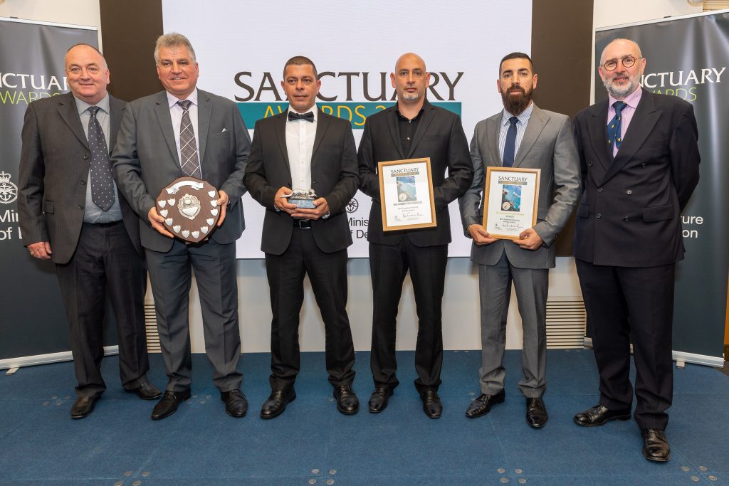Pictured are the Bird Steering Group from Cyprus who are a group of 6 men. They are wearing suits and ties. The individual second to the left is holding an award shaped like a shield and is wooden. It has silver little shields around it. Next to him on the right is an individual holding a Silver Otter Award shaped as a silver otter. Next to this individual are two individuals on the right holding wooden framed certificates.
