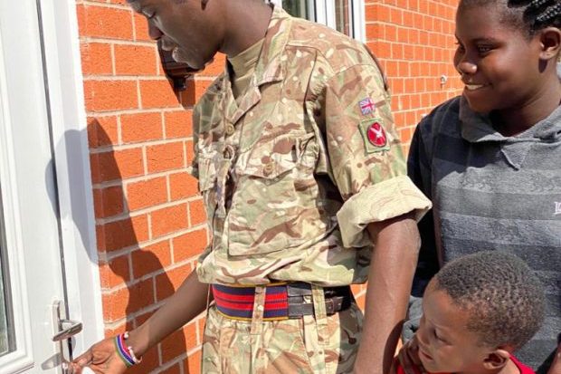 A British Army soldier from Kenya wearing his uniform is opening the door of his new Service Family Accomodation. Behind him is his daughter in a long-sleeved grey top and his small son in a red top.