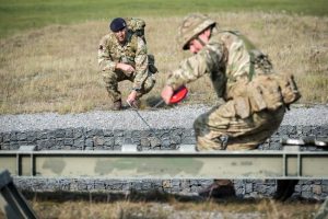 Two reservists are building an infantry assualt bridge out of grey rocks. They are standing opporsite each other and one is holding a rope to put the bridge together.