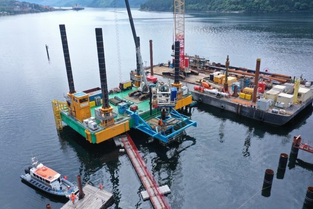 Pictured is the construction site of the Glen Mallan Jetty on the sea. It is square with two black cylinders on it and a construction crane. On the left is a small white boat and pictured behind the jetty are mountains.