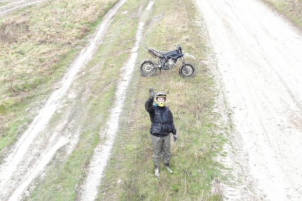 A dismounted motorbike rider waves at the camera above him. Behind him is his motorbike, just off the side of a track. 