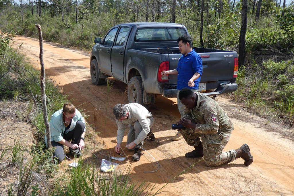 A blue pick up truck is halted on a dirt road. At the rear of it stands a man in a blue polo shirt. In front of him are three people kneeling - a man and a woman in civilian clothing and a man in military uniform. They are working on what appears to be the camera.
