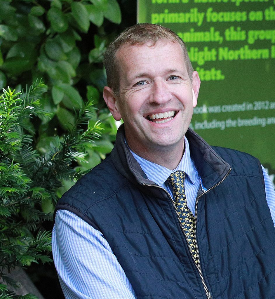 Alyn is a white man smiling at the camera. Behind him is some shrubbery and a green sign. He wears a blue shirt and tie with a navy gilet over the top.