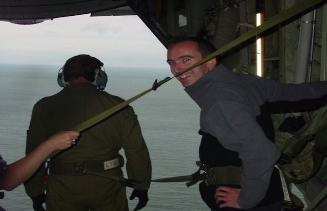 Warren is standing in a Hercules C-130J helicopter. Behind him the door is open, with an individual in headphones standing by him and the view of the sea.
