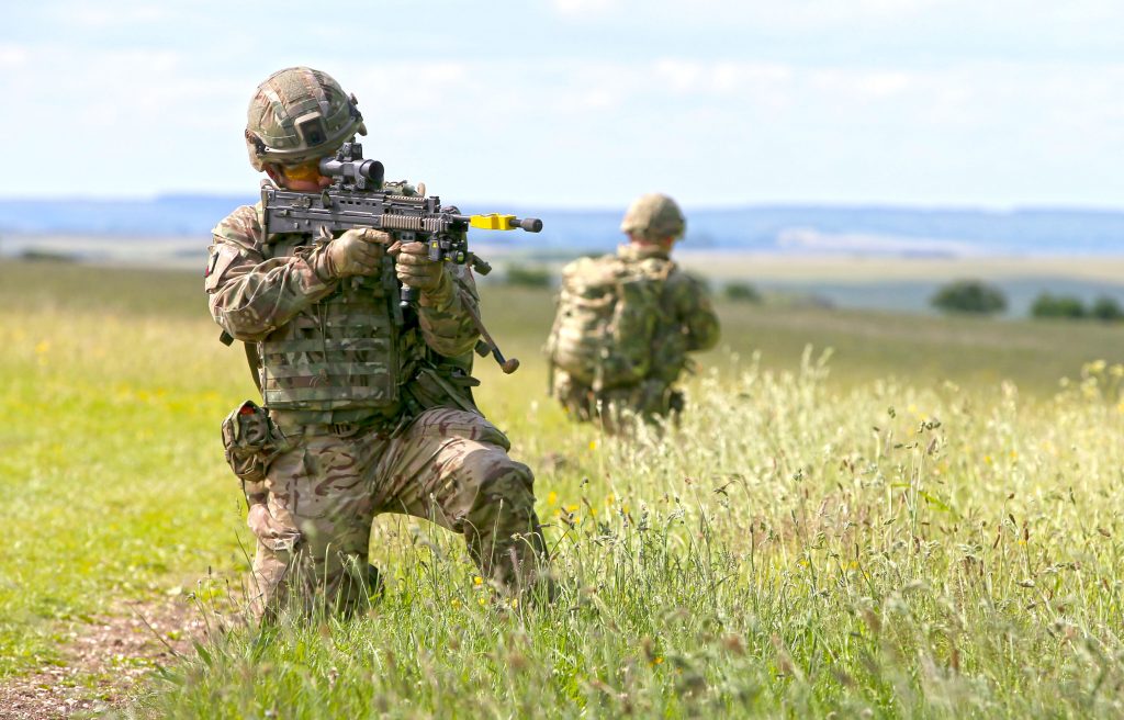A soldier is facing the front in armed gear and is pointing a gun in the distance. Behind him is another soldier facing towards the fields. Around them is greenery grass.