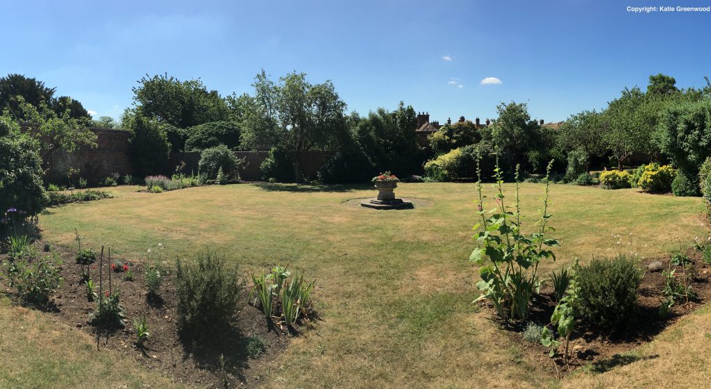 A large neat garden with flowerbeds on the left and right hand side, trees at the back and a fountain with flowers in the middle.