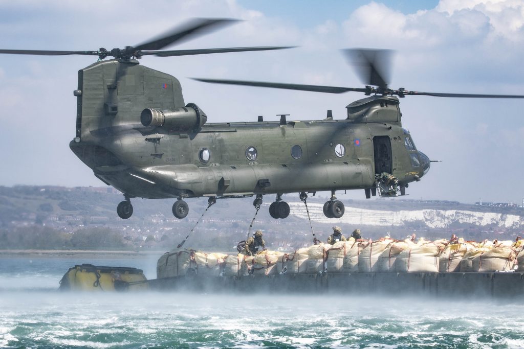 A Chinook hovers over a barge holding large bags of aggregate.