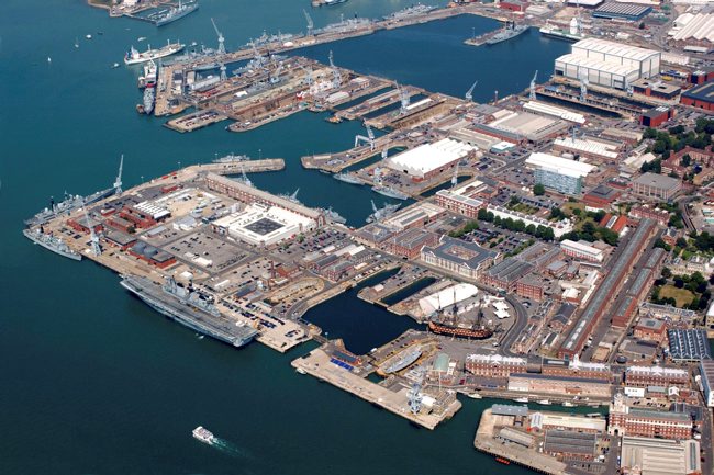 An aerial image of the Portsmouth Dockyard. There is a range of buildings around the sea as well as ships.