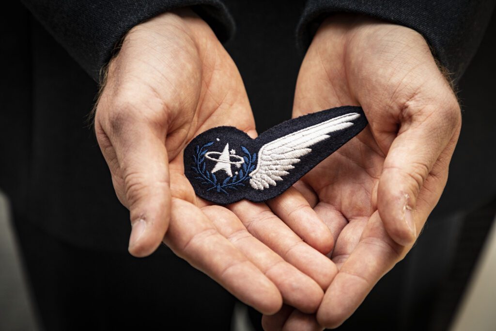 Two cupped hands hold a UK Space Command cloth badge. It has a dark background with an emblem on the left side. To the right is a single white wing.