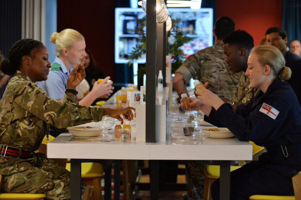 A group of male and female Service personnel are seated along a dining table, with plates of food and refreshments in front of them. They are conversing while they eat.