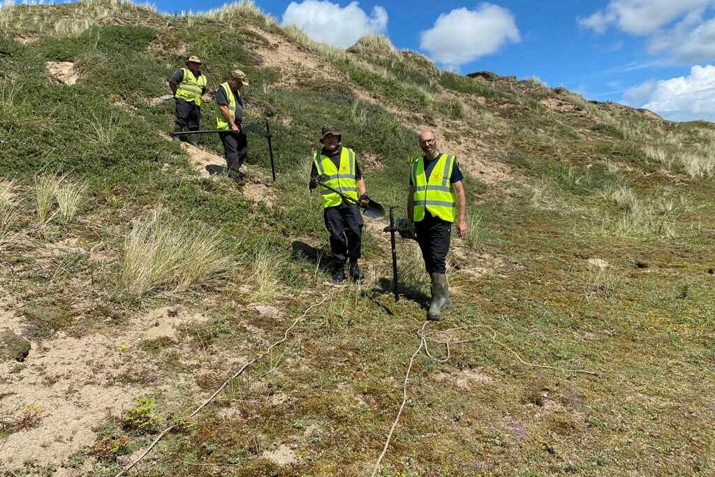 The photo shows four men from the explosive ordnance clearance team. They are walking towards the camera over an area of grassy dunes. They are wearing high-vis vests, and are carrying spades and metal detector equipment.