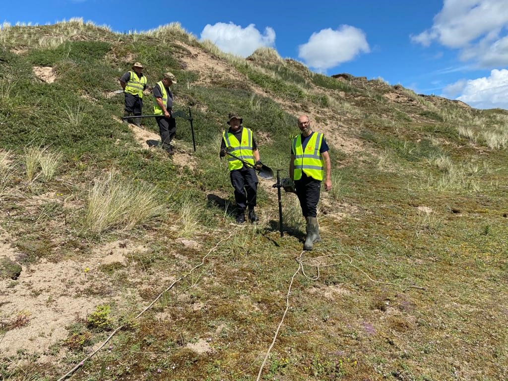 The photo shows four men from the explosive ordnance clearance team. They are walking towards the camera over an area of grassy dunes. They are wearing high-vis vests, and are carrying spades and metal detector equipment. 