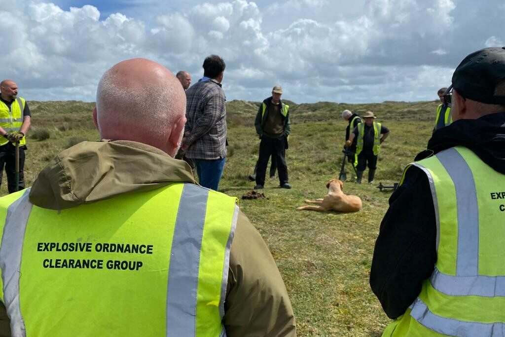 A group of eight men are stood in a wide circle on an area of level grass. They are wearing green high-vis jackets. There is a man in plain clothes and a dog in the middle of the circle. The man in the middle is giving a briefing to the EOD clearance personnel in the high-vis jackets.