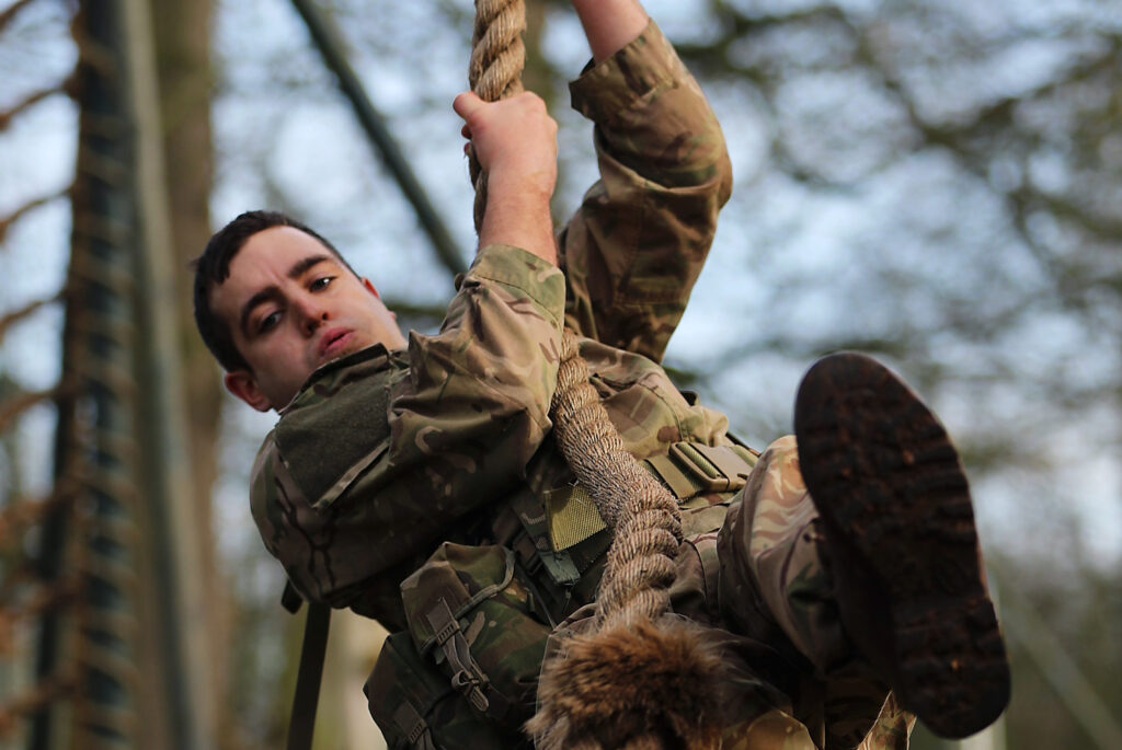 A Royal Marine recruit in uniform looks directly at the camera as he hangs from a rope. Both of his hangs grip the rope with his right leg curled under him and his left leg held out horizontally towards the camera. 