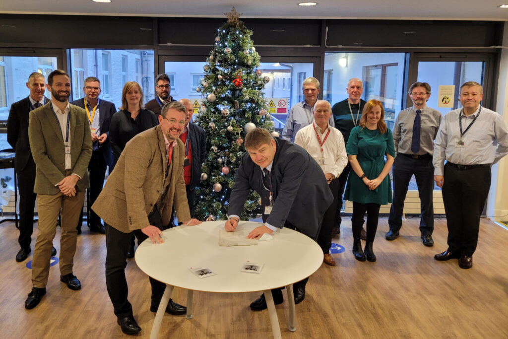A group of people grouped around a Christmas tree. In front of the larger group is a table with two men leaning over it, signing a document.