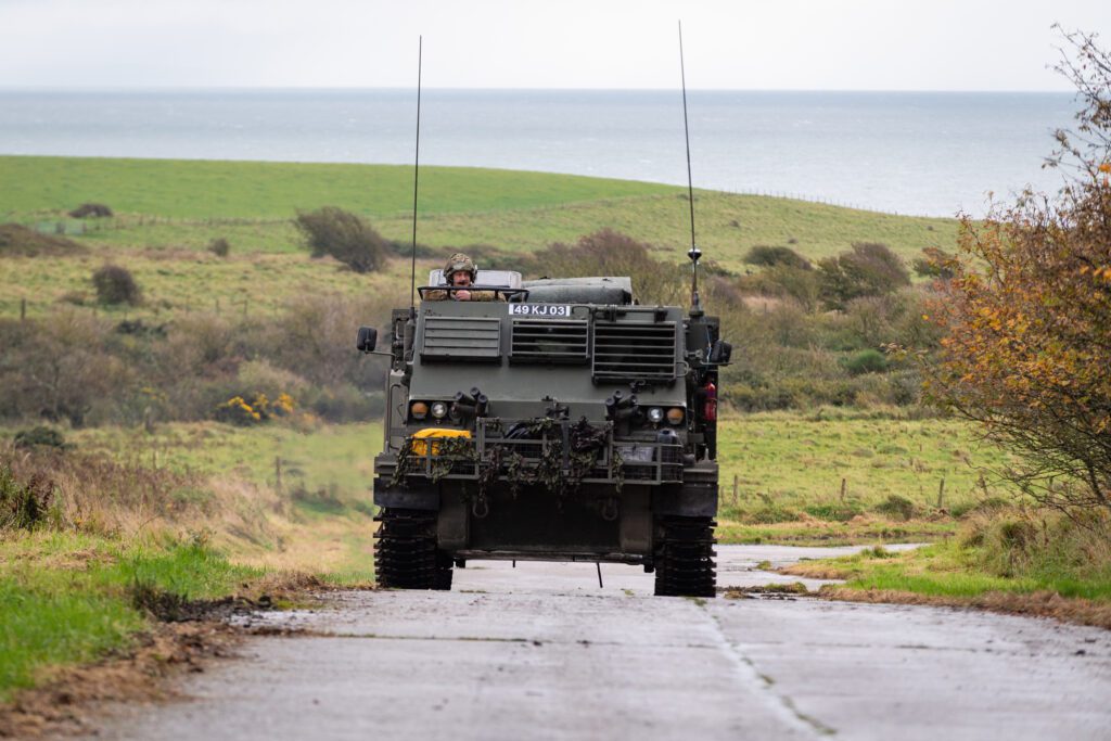 A large dark green vehicle drives along the road towards the camera. The head of a soldier is visible sticking up from the top of the vehicle. The background is green and open with the sea visible.