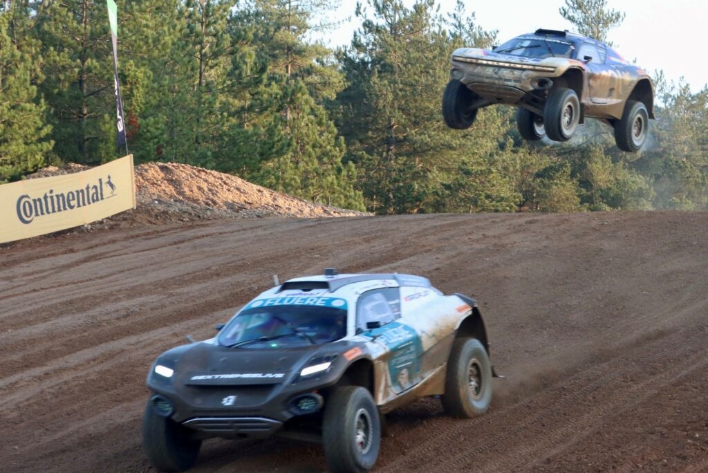 The photo was taken from the side of the racetrack and shows two Extreme E SUVs passing over a jump. The car in front is on the descent while the other follows closely behind in mid-air over the jump. In the background is a row of trees adjacent to the track.