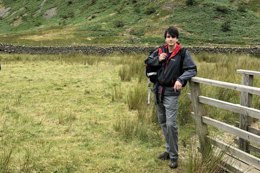 A white man wearing hiking gear and carrying a rucksack stands in an open field next to a wooden fence. In the background there is a dry stone wall with hills behind.