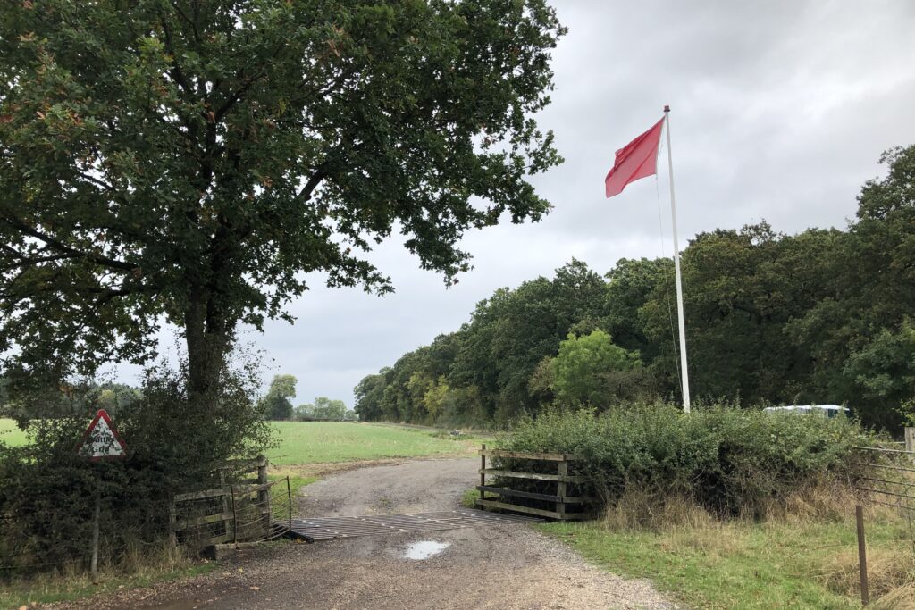 A red flag is flying atop a large white flagpole, next to a gravel path with a cattle grid in the centre of the photo. On the opposite side to the flag is a large tree, behind which is an area of open fields and trees.
