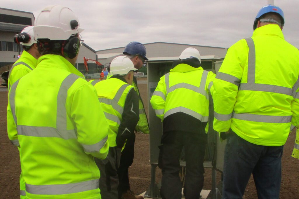 A group of men in high vis jackets and helmets lean in to look at a metal structure. 