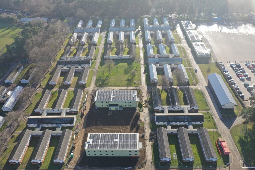 An aerial view of the two new double storey, carbon neutral accommodation buildings at Brunswick Camp. The buildings are a light green colour, with rows of solar panels along their roofs. Surrounding the two new buildings are a range of older barracks buildings and other facilities, with roads connecting the different areas of the camp.