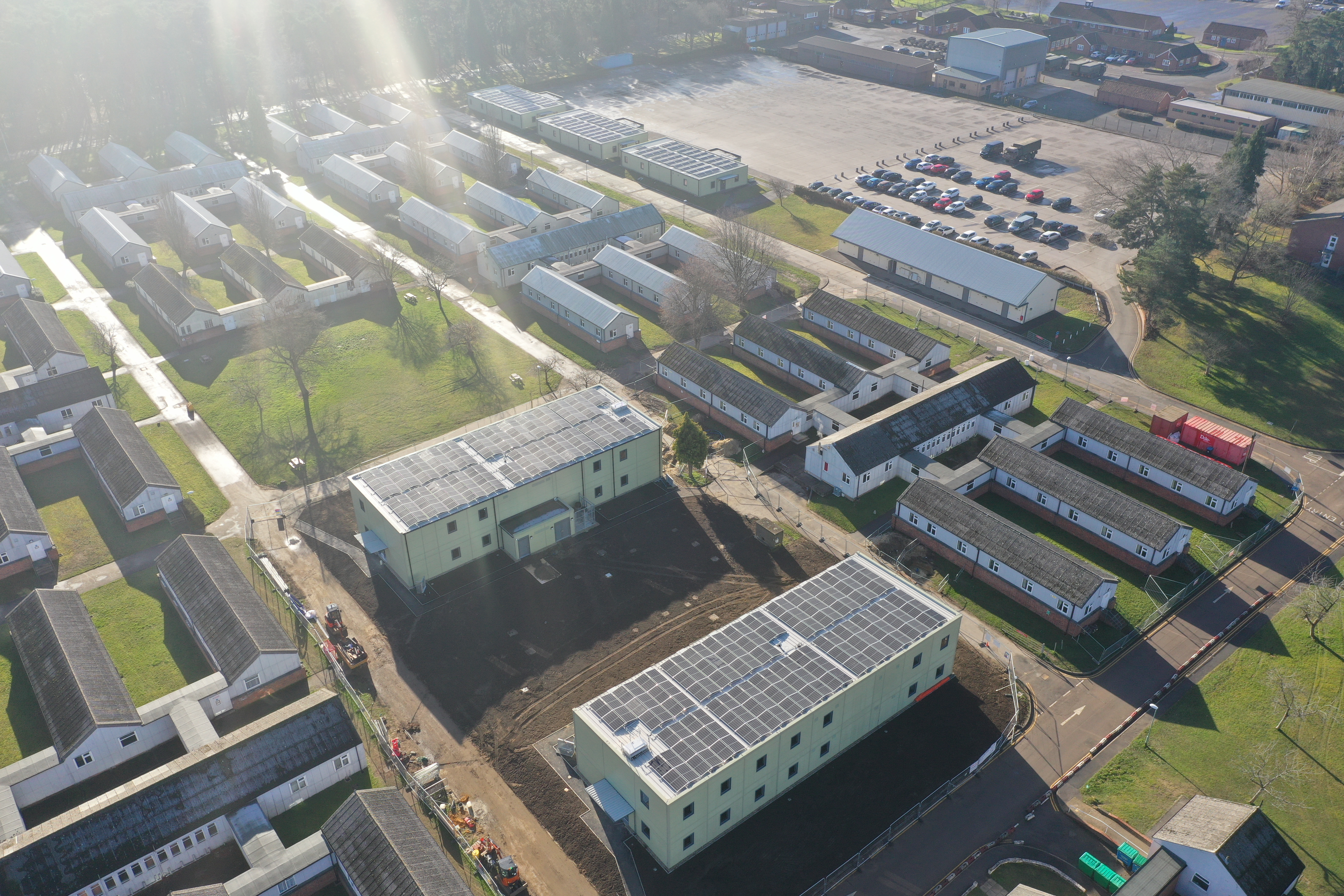 An aerial view of the two new double storey, carbon neutral accommodation buildings at Brunswick Camp. The buildings are a light green colour, with rows of solar panels along their roofs. Surrounding the two new buildings are a range of older barracks buildings and other facilities, with roads connecting the different areas of the camp.