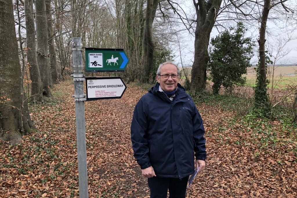 A white man stands in a leafy field with trees on either side. Next to him is a signpost marking a permissive bridleway and path for horse riders.