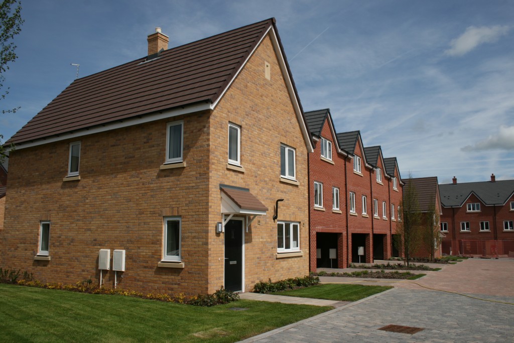 The photo shows a row of houses, fronted by grassy lawns and connected by a road which can be partially seen in the bottom corner of the image.