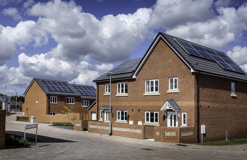 The photo shows a number of houses with solar panels on their roofs. In front of the houses is a paved, concrete area, with a lamp post and low brick walls between it and the houses.