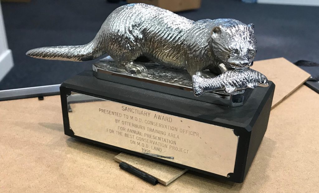 A trophy which features a silver, metal otter with a fish in its mouth, perched atop a black square base. On the base of the trophy is a plaque which reads: 'Sanctuary Award, presented to MOD conservation officer by Otterburn Training Area, for annual presentation for the best conservation project on MOD land, 1991.