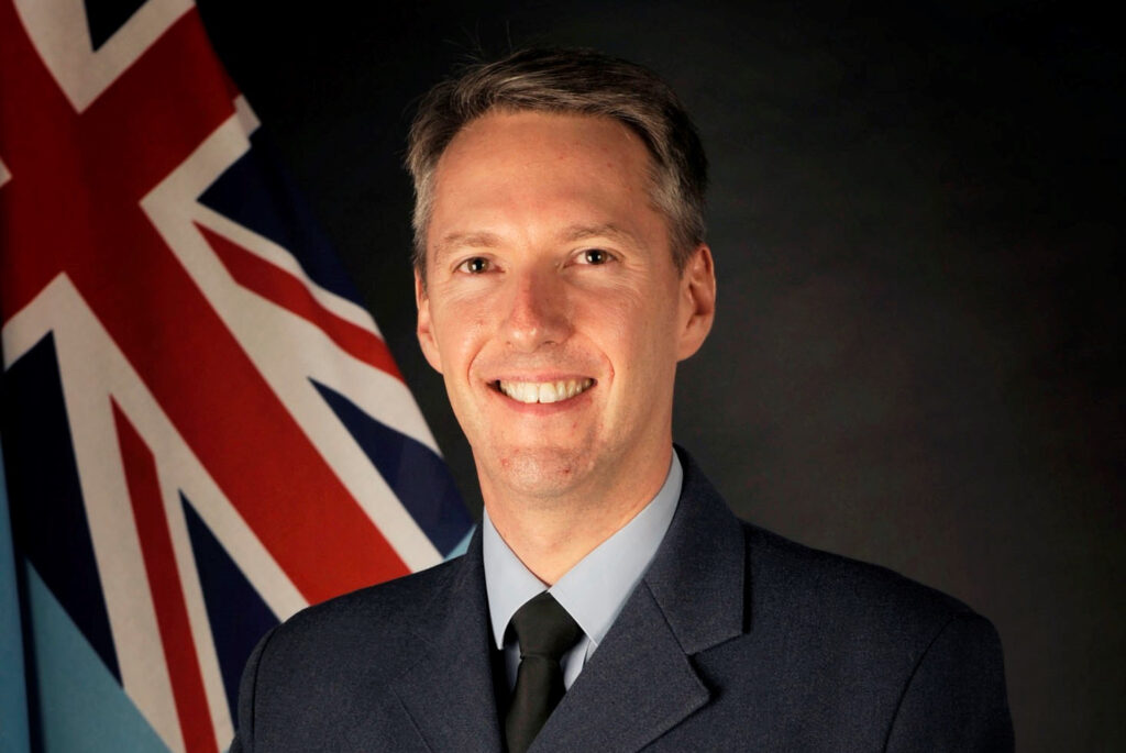 Air Commodore James Savage stands in a formal posed image. It shows from the waist and above. He is smiling at the camera, wearing RAF uniform with an RAF flag behind him.