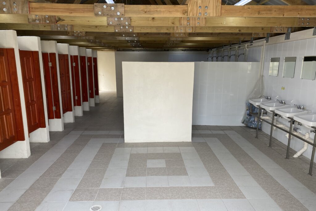 The interior of a building containing toilet cubicles, showers and sinks. The floor and walls are finished with clean, white and grey tiles, and the cubicle doors are made of wood. The ceiling has also has wooden rafters.