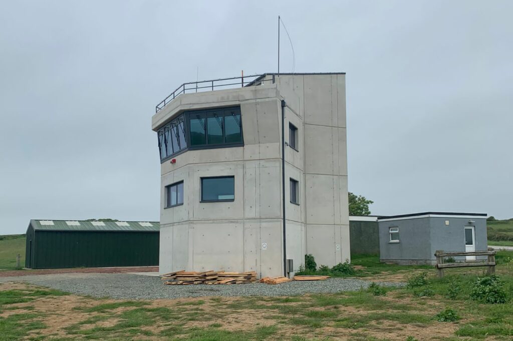 A view of the new tower. It appears to be a three storey concrete building. The ground floor does not have windows. The second floor has several and the top floor has a continuous line of windows. There are small portakabin style buildings on either side.
