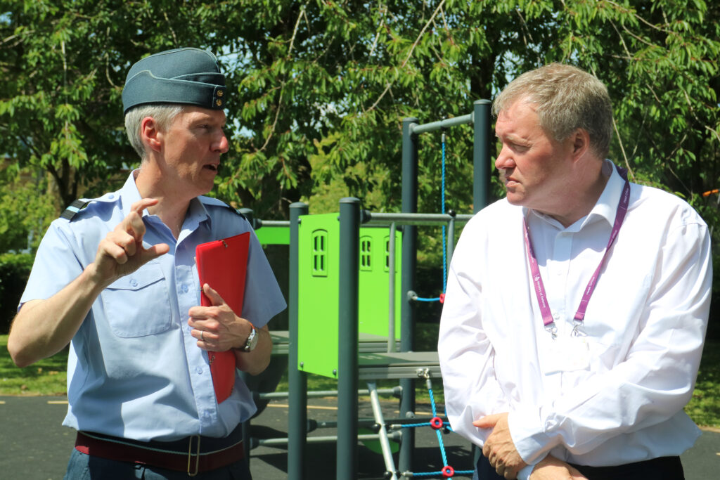Mike, wearing a white shirt, listens to and Air Commodore James Savage, wearing RAF uniform. They are outside and part of a children's play area is visible behind them.