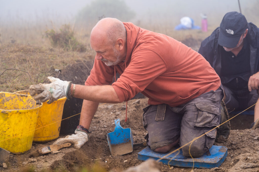 Two men kneel on the ground next to a shallow trench. One, wearing a light red hoodie, is placing an item into a bucket. The background is foggy.