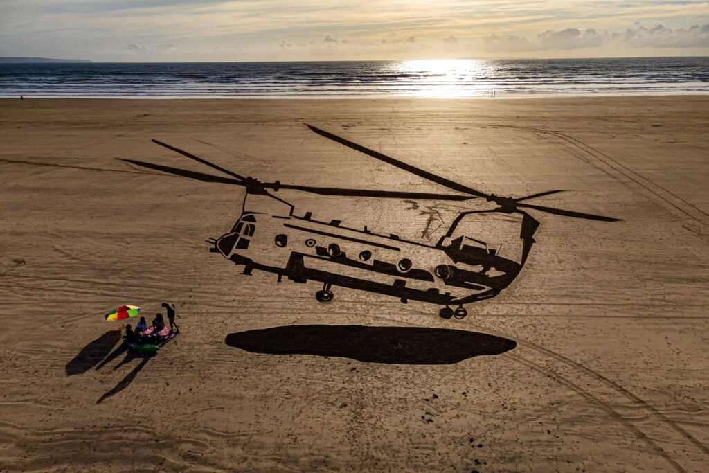 A view from above of a stretch of beach, into which a large sand drawing of a Chinook military helicopter has been drawn. Next to the Chinook artwork, as if below the helicopter, there is a small group of people looking as though they are having a picnic.
